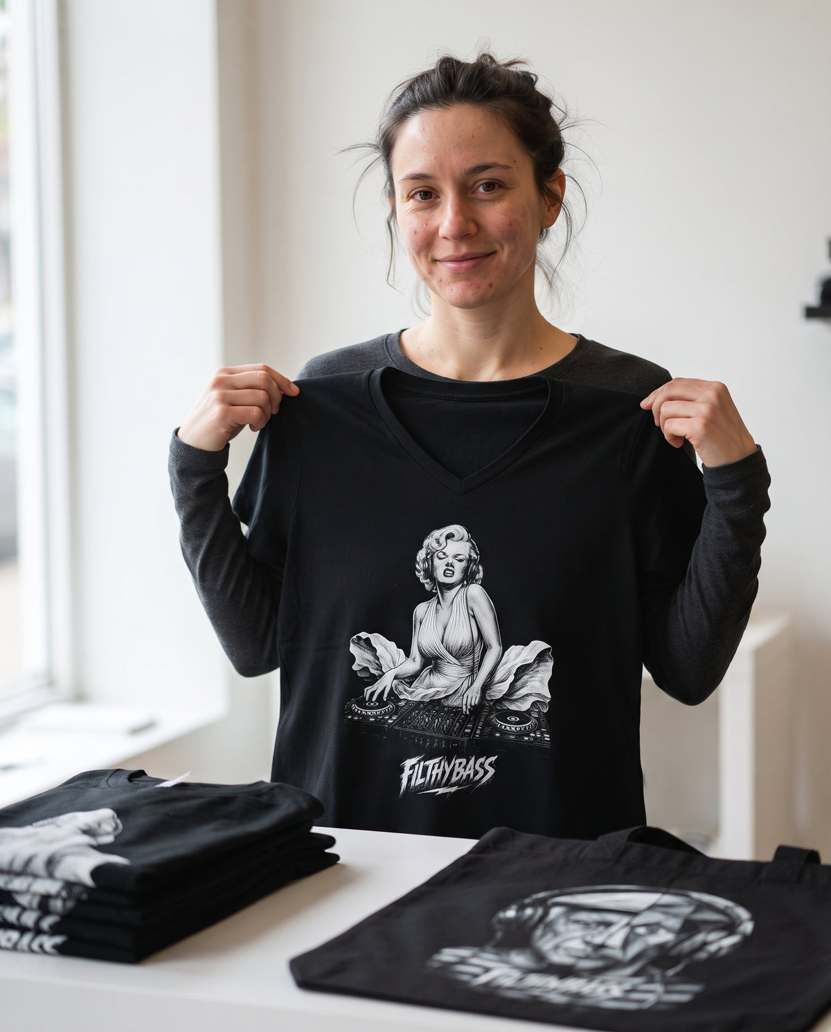 A person holding up a black FILTHYBASS V-neck t-shirt behind a table of folded stock