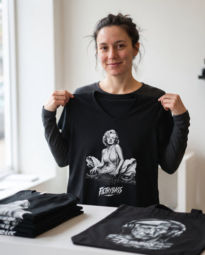 A person holding up a black FILTHYBASS V-neck t-shirt behind a table of folded stock